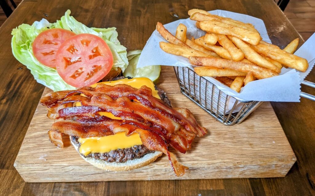 A burger with bacon and fries on a wooden cutting board.